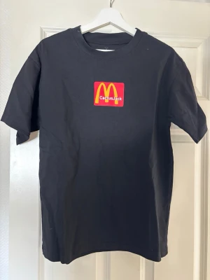 Svart Cactus Jack x McDonald's t-shirt - Säljer en svart t-shirt med Cactus Jack x McDonald's tryck framtill och ett unikt mönster på ryggen. T-shirten har rund hals och korta ärmar. Tillverkad i mjuk bomull, perfekt för dig som gillar streetwear och samarbeten.