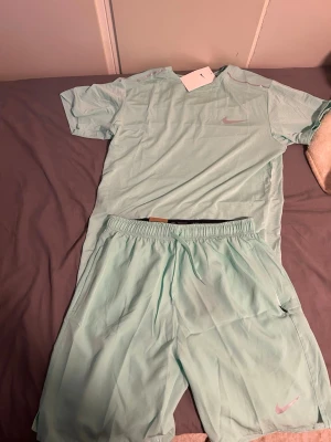 Mintgrönt träningsset från Nike - Säljer ett fräscht mintgrönt träningsset från Nike med t-shirt och shorts. Båda plaggen har Nike-logga i ljusrosa och är gjorda i ett lätt, snabbtorkande material som passar perfekt för träning eller sportaktiviteter.