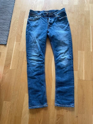 Blå raka jeans från Nudie Jeans - Säljer ett par klassiska blå jeans från Nudie Jeans med raka ben och knappgylf. Jeansen har snygga slitningar och kontrastsömmar. Perfekta till vardags för en avslappnad och stilren look.