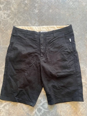 Svarta shorts från Valient - Snygga svarta shorts från Valient i klassisk modell med normal passform. Shortsen har bälteshällor, sidofickor och stängs med knapp och dragkedja. Perfekta för sommardagar och enkla att matcha med olika stilar.