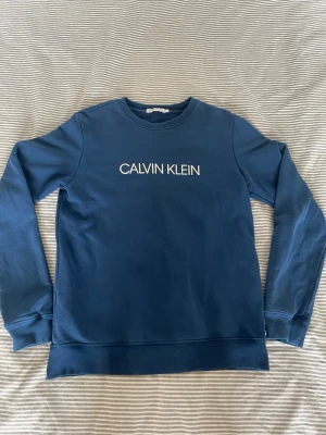 Blå sweatshirt från Calvin Klein - Snygg blå sweatshirt från Calvin Klein med vit logga framtill och en liten patch med texten 'JEANS' bak. Tröjan har rund hals, långa ärmar och ribbade muddar vid ärmslut och nederkant. Perfekt för en avslappnad och trendig look.