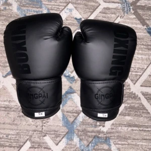 Svarta boxningshandskar GINGPAI 12 oz - Säljer ett par svarta boxningshandskar från GINGPAI i storlek 12 oz. Handskarna har en stilren design med texten 'BOXING' på ovansidan och bred kardborrestängning vid handleden för extra stöd. Perfekta för träning eller sparring.