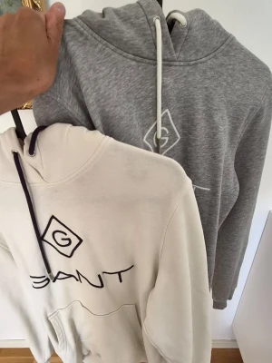 Två GANT hoodies med logga s/xs - Säljer två snygga hoodies från GANT, en i ljusgrått och en i offwhite. Båda har huva med dragsko, stor broderad GANT-logga på bröstet och känguruficka framtill. Perfekta för en avslappnad och trendig stil. 250kr för båda. Storlek S. Tycker nog att det är närmare xs. 