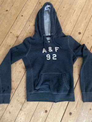 Mörkblå hoodie med A&F-tryck - Mörkblå hoodie med huva och magficka. På bröstet står det A&F 92 i vita bokstäver. Hoodien har långa ärmar och vita sömmar som kontrast. Perfekt för en avslappnad stil och enkel att matcha med jeans eller mjukisbyxor.pris kan diskuteras 