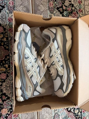 Asics gel nyc  - Snygga sneakers från Asics i vitt och grått med mesh- och syntetpaneler. Skorna har en chunky sula och klassisk Asics-logga på sidan. Perfekta för dig som gillar sportig och trendig stil. Lågt skaft och snörning framtill.