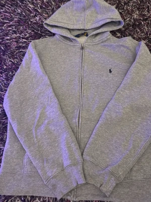 Grå zip hoodie från Polo Ralph Lauren - Säljer en stilren grå zip hoodie från Polo Ralph Lauren med klassisk logga broderad på bröstet. Hoodien har fickor framtill och ribbade muddar vid ärmar och nederkant. Perfekt för en avslappnad look och enkel att matcha med det mesta.          Pris går att diskutera vid snabb affär 