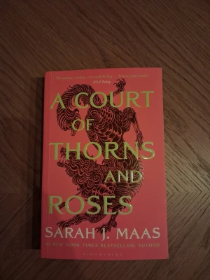 A Court of Thorns and Roses - En fantasyroman av Sarah J. Maas med ett rött och guldigt omslag och ett konstnärligt lejonmotiv. Boken handlar om Feyre som tas till fånga av en High Fae Lord och dras in i en magisk och farlig värld fylld av romantik och äventyr.