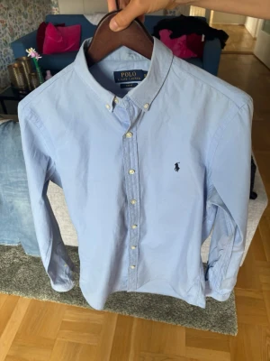 Ljusblå skjorta från Polo Ralph Lauren - Klassisk ljusblå skjorta från Polo Ralph Lauren med button-down krage och broderad logga på bröstet. Skjortan har långa ärmar, vita knappar och är tillverkad i bomull. Perfekt för en stilren och avslappnad look.
