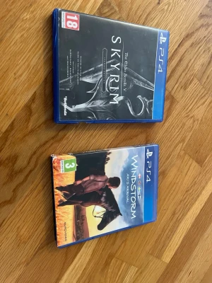 PS4 spel Skyrim & Windstorm - Säljer två PS4-spel: The Elder Scrolls V: Skyrim Special Edition med svart omslag och fantasytema samt Windstorm: Ari's Arrival med färgstarkt omslag och hästtema. Båda spelen kommer i blå plastfodral och har PEGI-märkning.