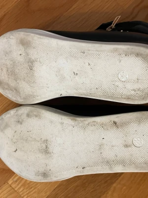 Svarta sneakers med nitar och dragkedja - Snygga svarta sneakers med vita sulor, dekorerade med små silverfärgade nitar på sidan och dragkedja i guld. Skorna har snörning och kardborreband upptill för extra stil och passform. Materialet ser ut att vara konstläder.
