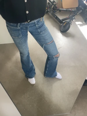 Blå slitna bootcut jeans - Säljer ett par blå bootcut jeans med slitningar och hål på ena benet. Jeansen har låg midja, klassiska fem fickor och en snygg tvättad look. Perfekta för dig som gillar en avslappnad och trendig stil. Väldigt lowwaist💕