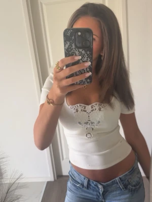 Vit croppad topp med spetsdetaljer - Säljer en vit croppad topp med korta ärmar och v-ringning. Toppen har fina spetsdetaljer framtill och sitter tight. Perfekt till jeans eller kjol för en trendig look.