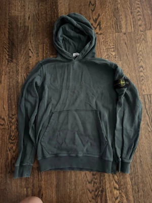 Mörkgrön hoodie med patch på ärmen - Snygg mörkgrön hoodie med huva och känguruficka framtill. Tröjan har en tydlig patch med logga på vänster ärm och ribbade muddar vid ärmslut och nederkant. Perfekt för en avslappnad streetwear-look.