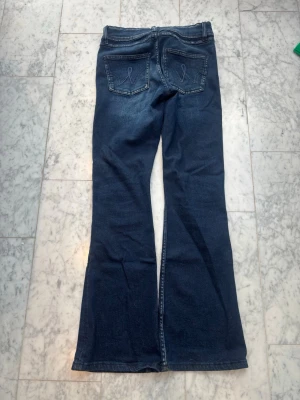 Mörkblå bootcut jeans med fickdetaljer - Säljer ett par mörkblå bootcut jeans med snygga fickdetaljer bak och klassisk femficksmodell. Jeansen har utsvängda ben och kontrastsömmar. Materialet är denim i bomull med lite stretch för extra komfort.