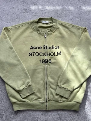 Grön zip hoodie Acne Studios - Snygg ljusgrön zip hoodie från Acne Studios med tryck 'Acne Studios STOCKHOLM 1996' på bröstet. Tröjan har ribbade muddar, rund halsringning och dragkedja framtill. Perfekt för en avslappnad streetwear-look.