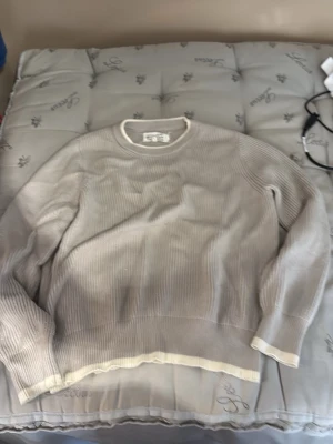 Ljusgrå stickad tröja Maison Margiela - Ljusgrå stickad tröja från Maison Margiela med ribbad struktur och kontrasterande vita kanter vid hals, ärmslut och nederkant. Tröjan har en avslappnad passform och rund halsringning. Perfekt för dig som gillar minimalistisk och stilren design.