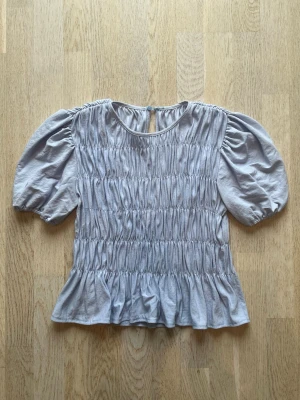 Ljusgrå blus med puffärm och volang - Supersöt ljusgrå blus med puffiga korta ärmar och volangkant nedtill. Smockad över hela livet för en snygg passform och rund halsringning med knapp i nacken. Perfekt att styla med jeans eller kjol. Tveka inte på att skriva en fråga eller be om fler bilder.🌸🌸