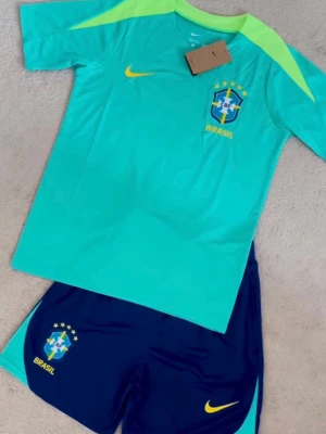 Brasilien träningsset Nike blå/grön - Säljer ett Brasilien träningsset från Nike med ljusgrön t-shirt och mörkblå shorts. Tröjan har gula detaljer och Brasilien-logga på bröstet, medan shortsen har matchande logga och gul Nike-swoosh. Perfekt för fotbollsträning eller sport.