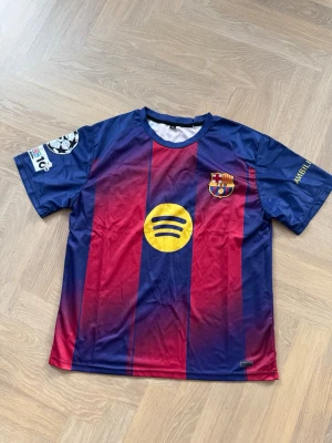FC Barcelona matchtröja Lamine Yamal - Säljer en officiell FC Barcelona matchtröja med Lamine Yamal och nummer 19 på ryggen. Tröjan har klassiska blå och röda ränder, klubbmärke på bröstet, Champions League-märke på ärmen och Spotify-logga på framsidan. Tillverkad i lätt och ventilerande material. Storlek XL men sitter som L