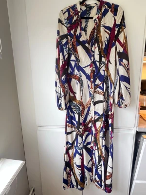 Mönstrad långklänning med knappar - Snygg långklänning med färgstarkt abstrakt mönster i blått, beige, lila och orange. Klänningen har långa ärmar, v-ringning och knappar framtill. Tråden på två knappar behöver fästas och när jag synar hittar jag en liten prick, går ev bort vid fläckborttagning men har ej testat. Inget som syns om man inte synar men har tagit bild på den för kännedom. Annars i mycket fint skick men därav priset och nivån jag satt.  