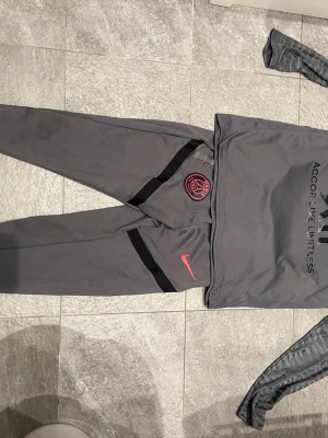 Gråa PSG träningsbyxor Nike - Säljer ett par gråa PSG träningsbyxor från Nike med rosa logga och klubbmärke. Byxorna har svarta detaljer och smal passform, perfekta för fotbollsträning eller gymmet. Elastisk midja för skön komfort.