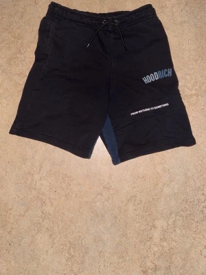 Svarta Hoodrich shorts  - Svarta shorts från Hoodrich med tryck på vänster ben där det står 'HOODRICH' och 'FROM NOTHING TO SOMETHING'. Shortsen har snörning i midjan och fickor på sidorna. Tillverkade i mjukt bomullsmaterial och har en avslappnad passform.