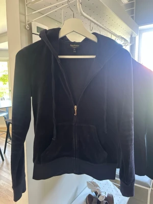 Mörkblå juicy couture hoodie med dragkedja - En juicy couture hoodie i mjuk velour med dragkedja framtill och stora fickor. Tröjan har huva med snören och ribbade muddar vid ärmslut och nederkant. Perfekt för en chill och avslappnad stil.