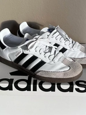 Adidas Samba vita och svarta sneakers - Klassiska Adidas Samba sneakers i vitt läder med svarta ränder och grå mockadetaljer på tån. Skorna har vit sula, vita skosnören och den ikoniska Adidas-loggan på plösen. Perfekta för en sportig och stilren look.
