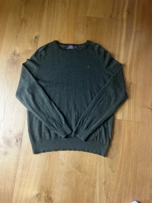 Mörkgrön stickad tröja Morris - 100% merino mörkgrön stickad tröja från Morris med rund halsringning och diskret broderad logga på bröstet. Tröjan har långa ärmar och ribbade muddar vid ärmslut och nederkant. Perfekt för lager-på-lager och stilrena outfits.