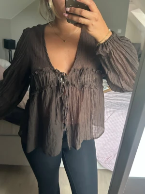 Brun transparent blus med knytning - Säljer en brun, transparent blus med djup v-ringning och knytning framtill. Blusen har ett lätt, skrynkligt tyg och långa, luftiga ärmar. Perfekt för dig som gillar en avslappnad men ändå trendig stil.