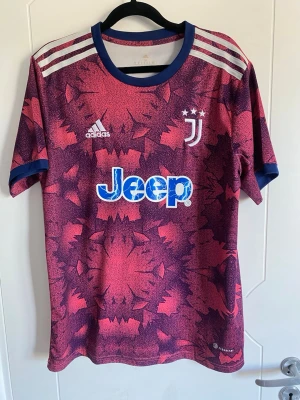 Juventus Adidas rosa matchtröja - Juventus matchtröja i rosa och mörklila med grafiskt mönster. Tröjan har blå detaljer vid krage och ärmslut, tre vita ränder på axlarna och tryckta loggor från Adidas, Juventus och Jeep. Perfekt för fotbollsträning eller som supporterplagg.