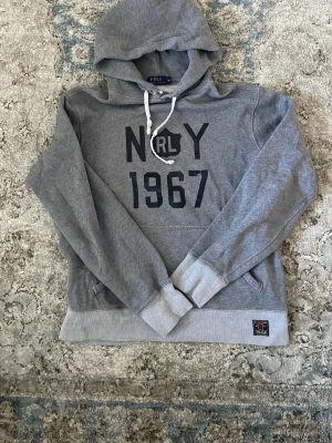 Grå hoodie Polo Ralph Lauren M - Snygg grå hoodie från Polo Ralph Lauren med trycket 'N R L 1967' på bröstet. Tröjan har huva med vita snören, ribbade muddar och en mjuk insida. Perfekt för en avslappnad och trendig look.