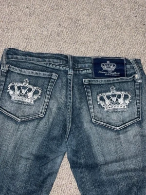 Blå Victoria Beckham jeans med brodyr - Säljer ett par blå jeans från Victoria Beckham med snygg slitning och broderade kronmotiv på bakfickorna. Jeansen har bootcut-modell och klassisk femficksdesign. Perfekta för dig som vill ha en unik detalj på dina jeans!