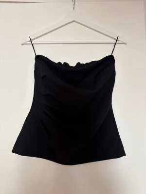 Svart axelbandslös tubtopp - Säljer en svarta axelbandslösa tubtopp med smala band och draperad framsida. Toppen har en enkel och stilren design som passar perfekt till många olika looks. Materialet känns mjukt och följsamt.