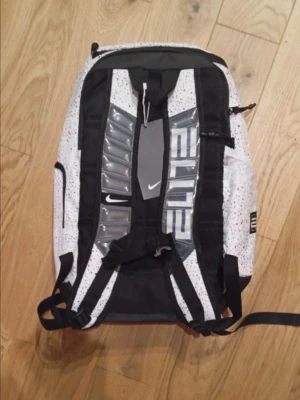 Vit och svart Nike Elite ryggsäck - Säljer en Nike Elite ryggsäck i vitt med svarta detaljer och färgstänk i rött och svart. Väskan har flera fack, vadderade axelband och tydlig ELITE-logga på sidan. Materialet är slitstark polyester och designen är sportig och modern.