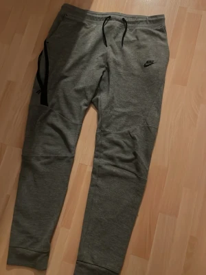 Gråa mjukisbyxor från Nike - Snygga gråa mjukisbyxor från Nike med svart logga på vänster lår. Byxorna har dragsko i midjan, muddar vid bensluten och sidofickor. Perfekta för chill eller träning, tillverkade i mjukt och bekvämt material.