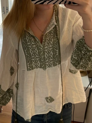 Vit blus med grönt mönster - Säljer en luftig vit blus med grönt broderat mönster och v-ringning. Blusen har långa ärmar med breda muddar och dekorativa detaljer. Perfekt för dig som gillar boho-stil och vill ha något unikt i garderoben.