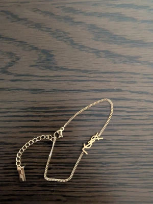 Guldarmband från Saint Laurent - Snyggt och stilrent guldarmband från Saint Laurent med den ikoniska YSL-logotypen i mitten. Armbandet har en tunn kedja och justerbar längd med en liten bricka där märket står ingraverat. Perfekt accessoar för att lyfta vilken outfit som helst.