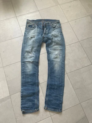 Blå slitna raka jeans - Säljer ett par blå jeans från nudie i modell LEAN DEAN med raka ben och slitningar på låren och knäna. Jeansen har använts och lagats tidigare men är i väldigt fint skick, små snygga slitningar på framsidan av jeansen och en väldigt snygg wash. Tveka inte att ställa frågor☺️