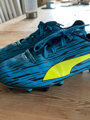Blåa fotbollsskor från Puma - Snygga blåa fotbollsskor från Puma med svart mönster med puma-loggan på sidan från puma i storlek 34. Skorna har svarta dobbar och svart insida. (Skorna har lite skråmor men är fina ändå, vill igentligen ha ”bra skick/acceptabelt skick”😅)💕Hör av er vid frågor eller funderingar🥰
