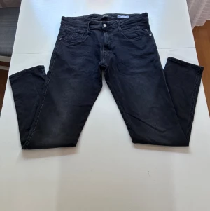 Svarta Replay Anbas - Säljer ett par svarta jeans med rak passform och klassisk femficksdesign. Jeansen har knappgylf och silverfärgade nitar. Materialet är denim med lätt tvättad look som ger en cool vibe. Perfekta till en avslappnad stil.