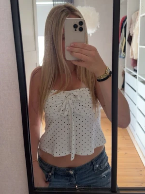 Vit prickig strapless topp med snörning - Söt vit topp med svarta prickar och snörning framtill. Toppen är ärmlös och croppad med en lätt volang nedtill💕