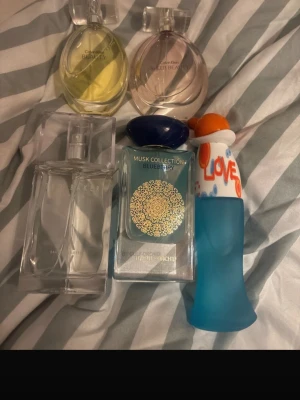 Mix av fem parfymer i olika färger - Säljer ett set med fem parfymer i olika flaskor och färger. Två flaskor är runda från Calvin Klein, en i gult och en i rosa. En rektangulär flaska från Ted Baker i genomskinligt glas, en blå flaska med LOVE-tryck och en blågrön flaska med guldmönster från Musk Collection Blueberry. Ca 80-90% kvar i varje 