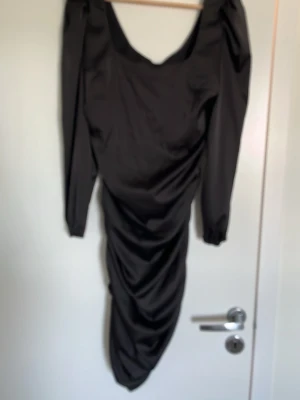 Svart långärmad draperad klänning - Snygg svart klänning med långa puffärmar och draperad effekt längs kjolen. Djup urringning framtill och figurnära passform ger en trendig siluett. Perfekt för dig som gillar stilrena och moderna plagg.