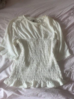 Vit topp med smock och volangkant - Säljer en söt vit topp med smockad midja och volangkant nedtill. Toppen har korta ärmar och rund halsringning. Perfekt för dig som gillar en feminin och trendig stil.