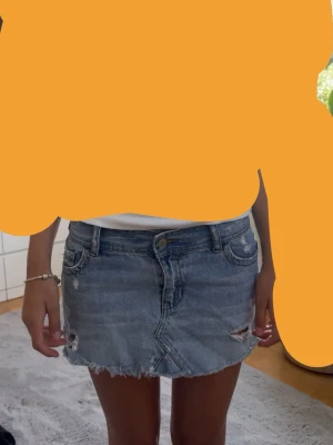 Kort jeanskjol med slitningar - Säljer en kort jeanskjol i blå denim med slitna detaljer och fransig nederkant. Kjolen har klassiska fickor fram och bak samt bälteshällor. Perfekt för dig som gillar en avslappnad och trendig stil.