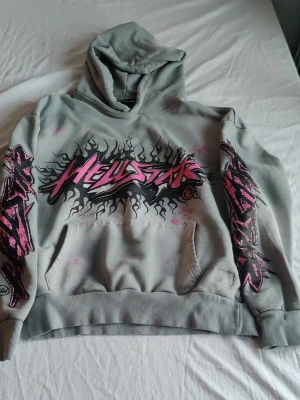 Grå Hellstar hoodie med tryck - Säljer en grå hoodie från Hellstar med stor grafisk logga i rosa och svart på bröstet och ärmarna. Hoodien har huva och magficka, samt coola detaljer i form av rosa stänk och tryck längs båda ärmarna. Perfekt för dig som gillar streetwear och vill sticka ut.
