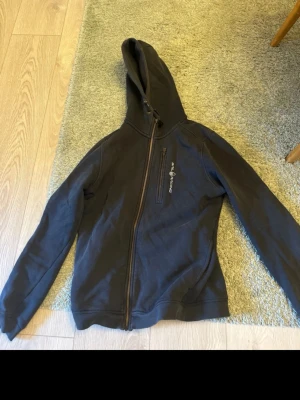 Svart hoodie från Sail Racing - Säljer en svart hoodie från Sail Racing med dragkedja och broderad logga på bröstet. Hoodien har två fickor framtill och är fodrad med mjukt material på insidan. Perfekt för dig som gillar sportig och stilren look.