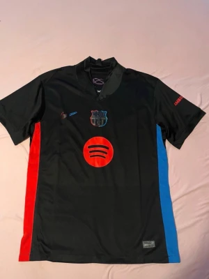 Svart FC Barcelona matchtröja - Snygg svart FC Barcelona matchtröja med röda och blå detaljer på sidorna. Tröjan har klubbmärke och Nike-logga på bröstet samt Spotify-logga i rött framtill. Tillverkad i lätt och ventilerande material, perfekt för fotboll eller träning.