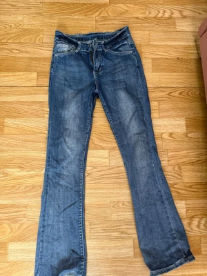 Blå bootcut jeans med slitningar - Säljer ett par blå bootcut jeans med snygga slitningar och tvättade detaljer. Jeansen har klassisk femficksdesign och dekorativa sömmar på bakfickorna. Materialet är denim i bomull och passformen är något utsvängd nedtill. Knappen har blivit lite lösare men går nog o fixa 